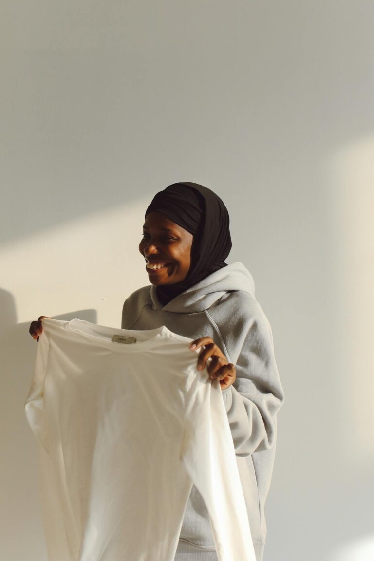 A cheerful woman holds up a white shirt while wearing a gray hoodie, standing indoors with warm lighting.