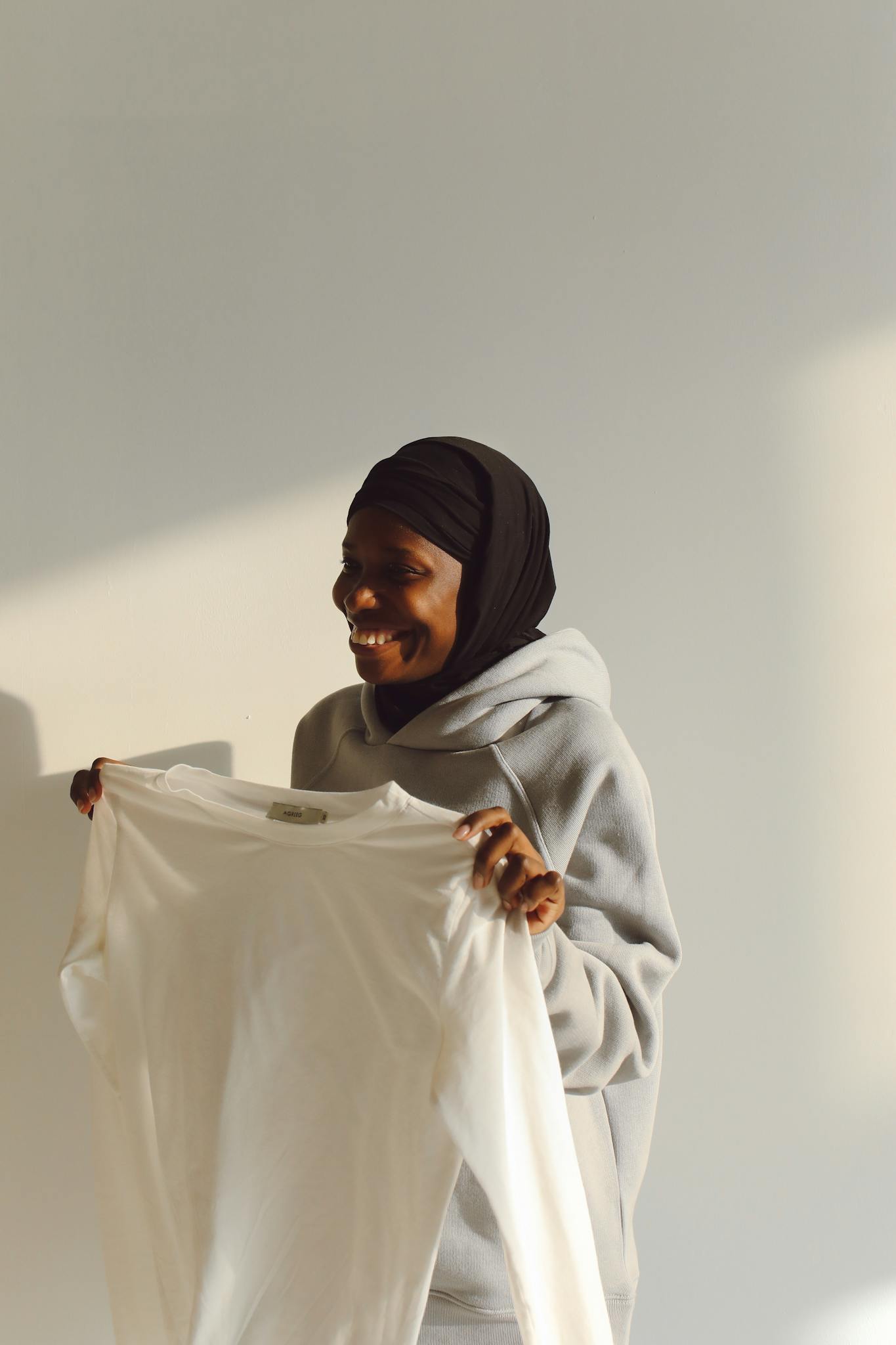 A cheerful woman holds up a white shirt while wearing a gray hoodie, standing indoors with warm lighting.